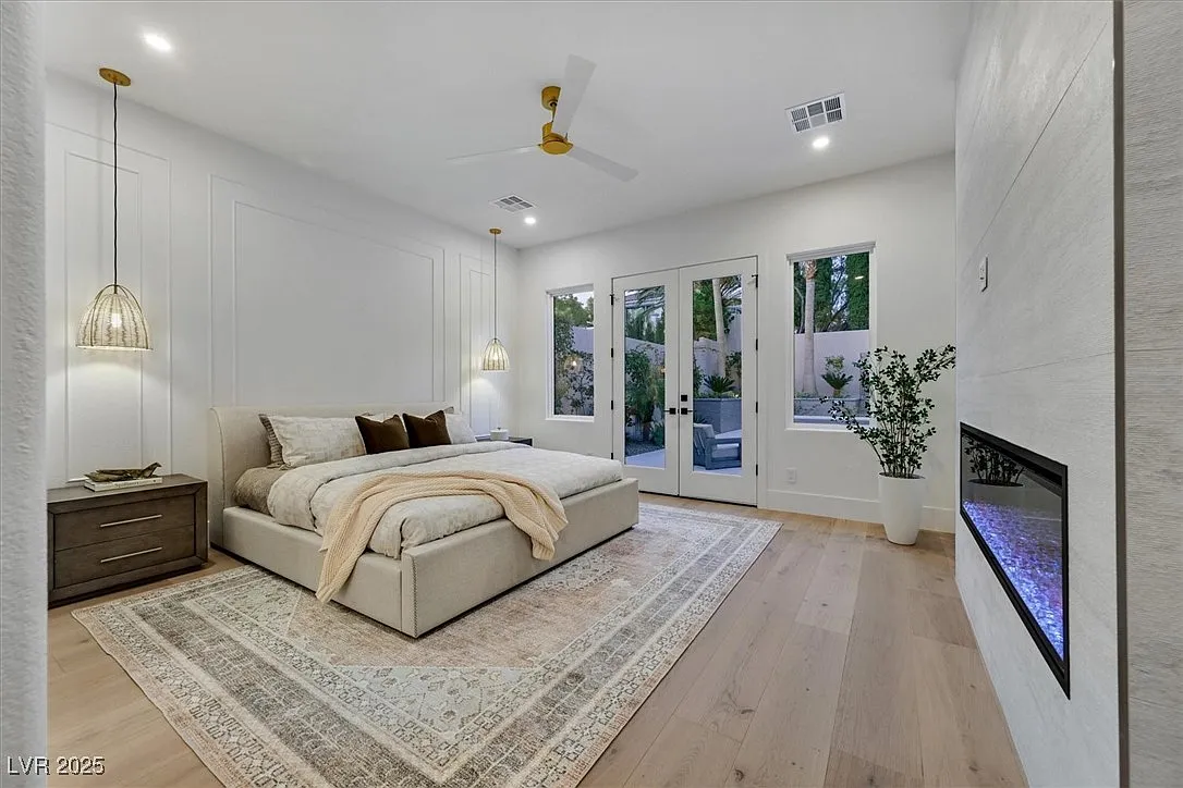 Contemporary primary bedroom with light wood floors, vaulted ceiling, pendant lighting, modern fireplace and French doors opening to a private patio.